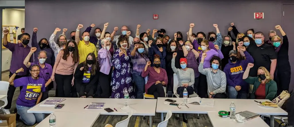 A large, diverse group of people pose indoors with raised fists, many wearing purple or SEIU shirts and face masks.