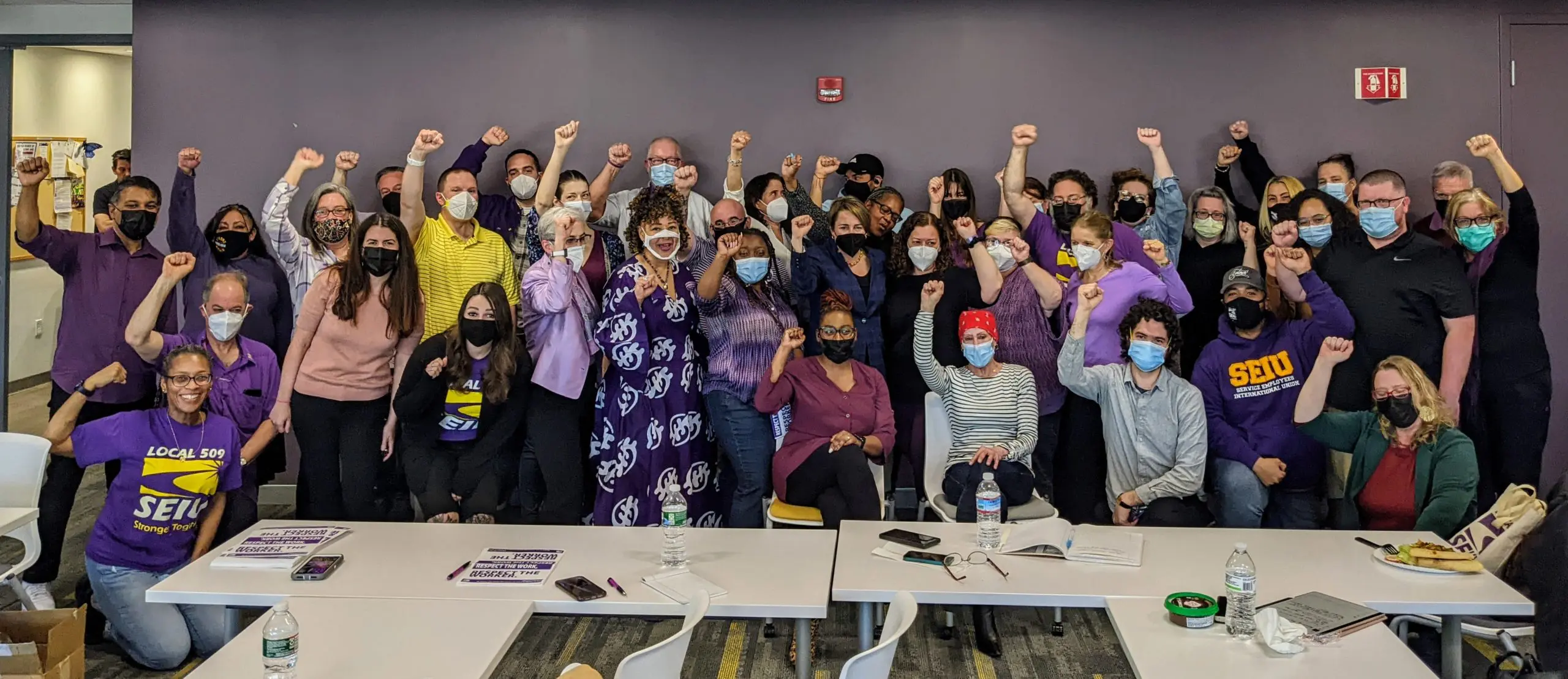 A large, diverse group of people pose indoors with raised fists, many wearing purple or SEIU shirts and face masks.