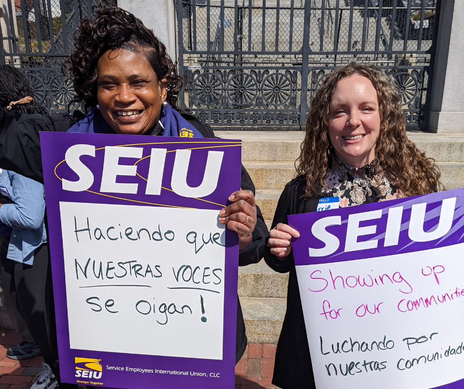 Two women smile and hold SEIU signs at a rally. One sign reads in Spanish, "Haciendo que nuestras voces se oigan!" The other in English and Spanish, "Showing up for our community. Luchando por nuestras comunidades.