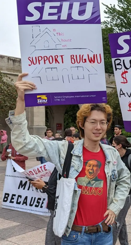 A person with dyed hair holds a sign reading "In this house we SUPPORT BUGWU" with a drawing of a house. Other people and signs are visible in the background.
