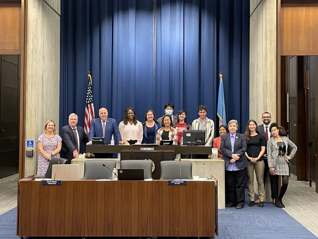 A group of people poses together in a government meeting room, standing behind and beside desks. An American flag and another flag are displayed behind them, with blue curtains as the backdrop.