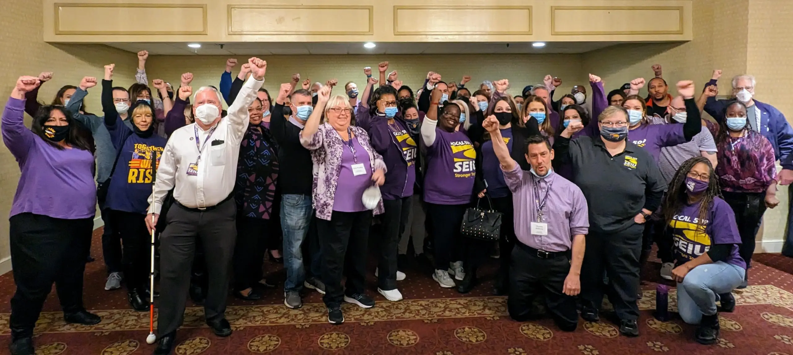 A large, diverse group of people, many wearing purple SEIU shirts, stand indoors with raised fists, showing solidarity.