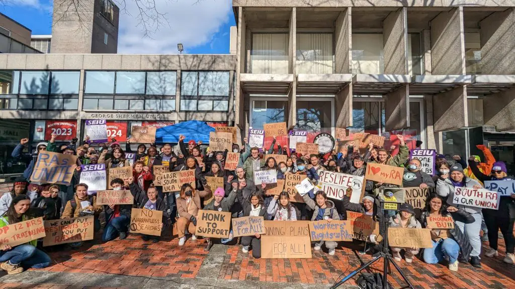 A large group of people pose together outside a building, holding handmade signs supporting RA unionization, workers' rights, and fair pay.