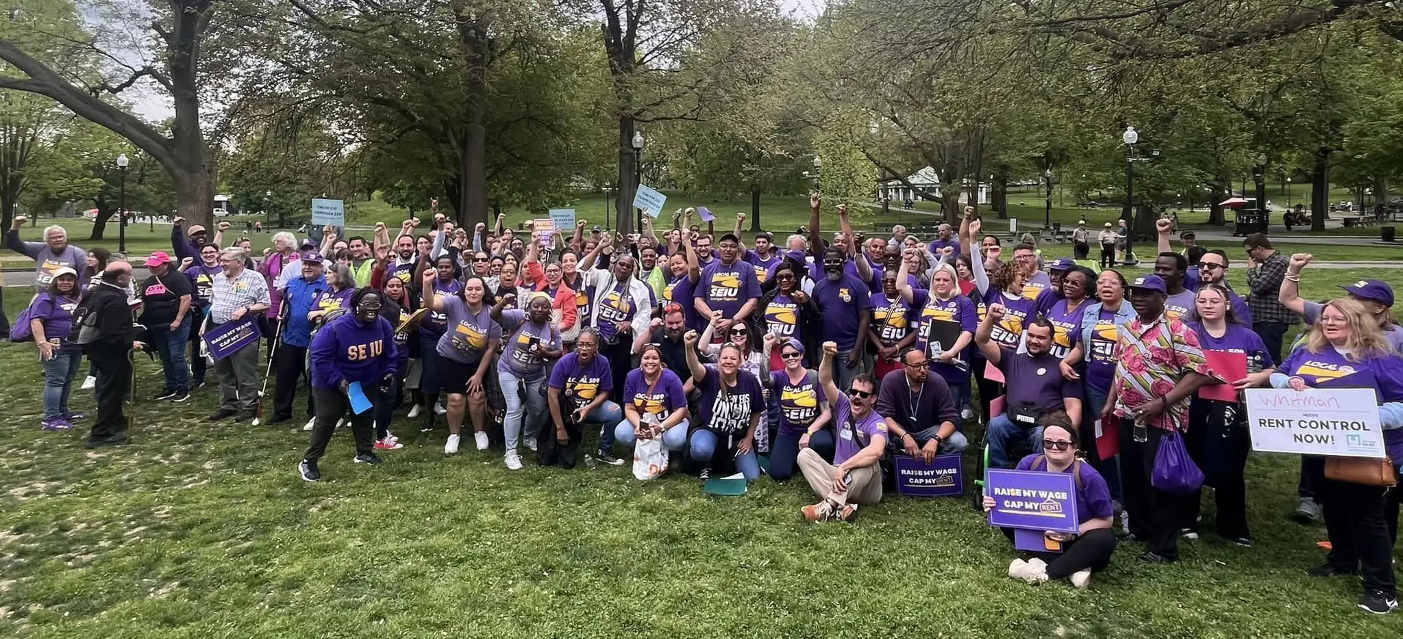 A large group of people wearing purple shirts and holding signs gather in a park, raising their fists in solidarity for rent control and fair wages. Trees and green grass are visible in the background.