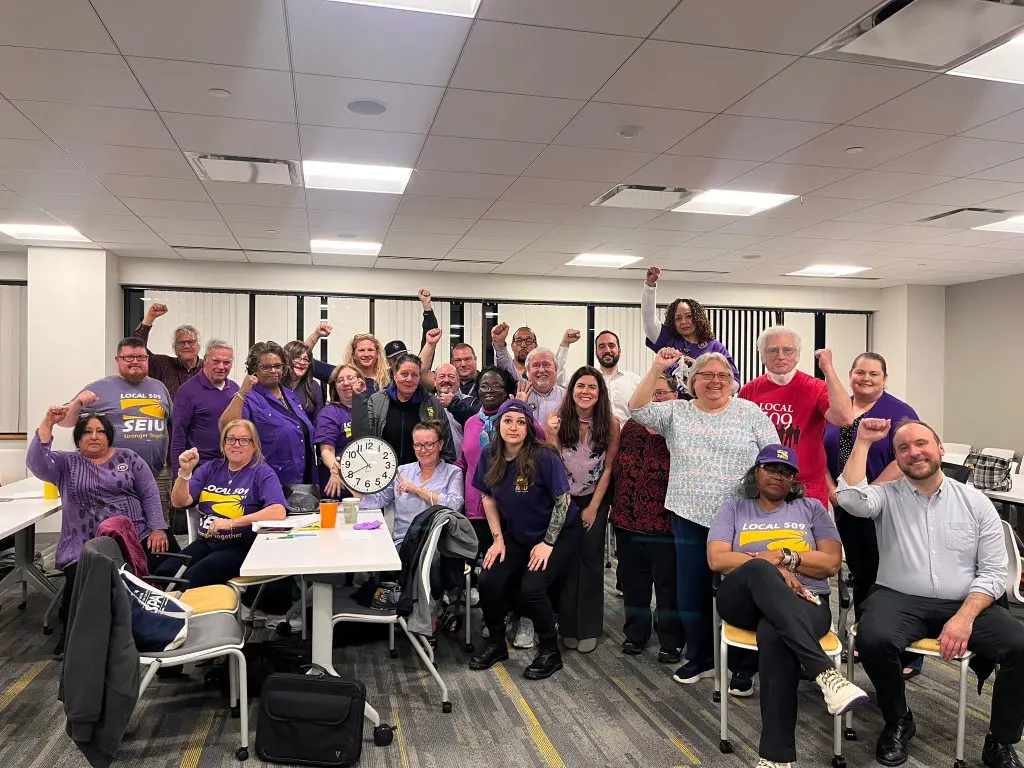 A group of people in a meeting room, many wearing purple shirts with "SEIU" logos, pose together smiling and raising their fists in solidarity.