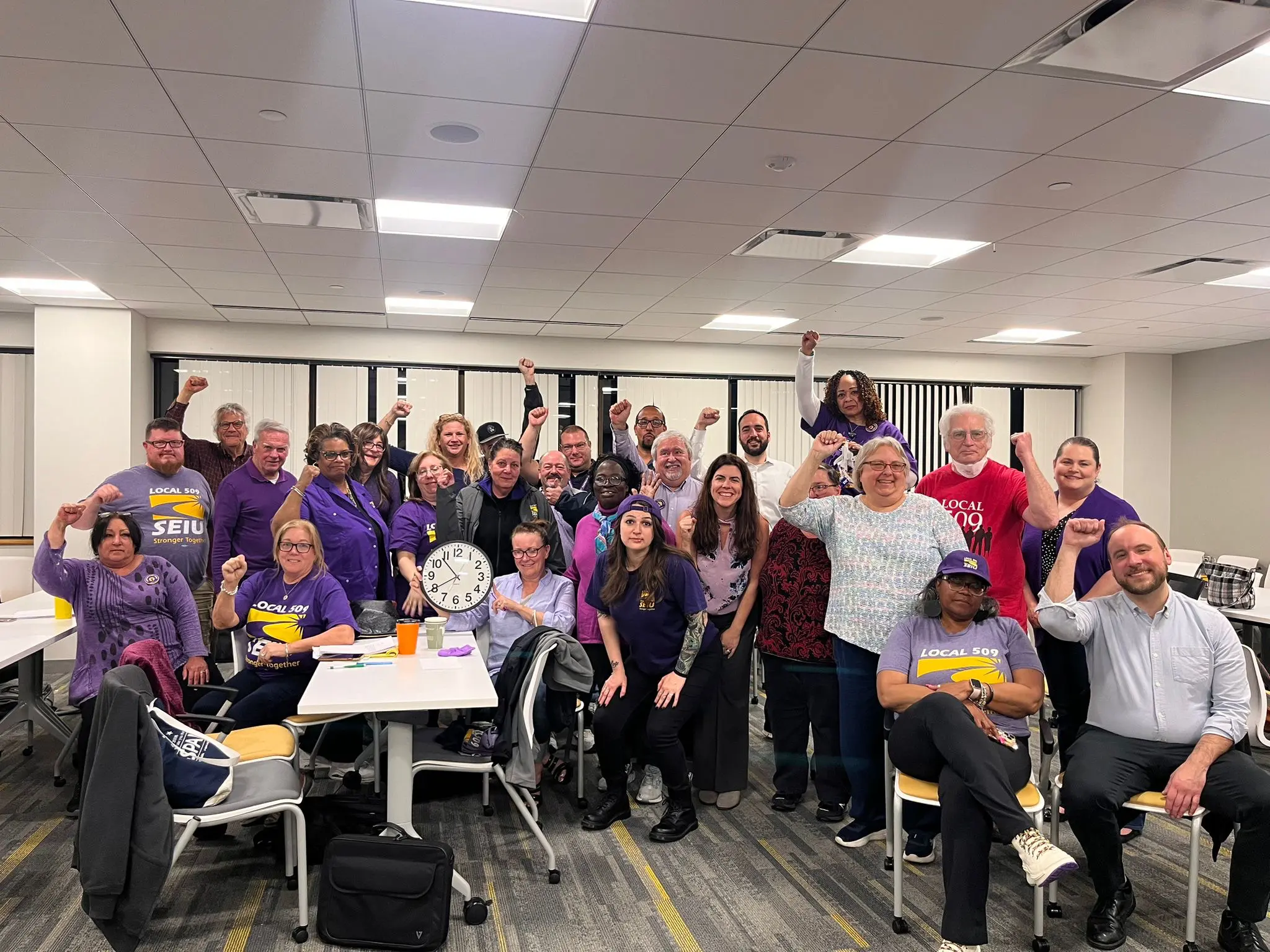 A group of people in a meeting room, many wearing purple shirts with "SEIU" logos, pose together smiling and raising their fists in solidarity.