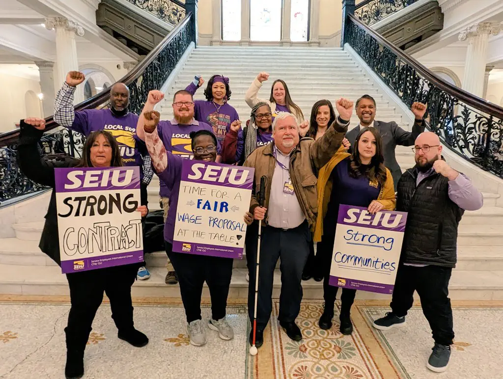 A diverse group of people stand on marble stairs, holding SEIU signs reading “STRONG CONTRACT,” “TIME FOR A FAIR WAGE PROPOSAL AT THE TABLE,” and “STRONG COMMUNITIES.” Most wear purple shirts and raise fists in solidarity.