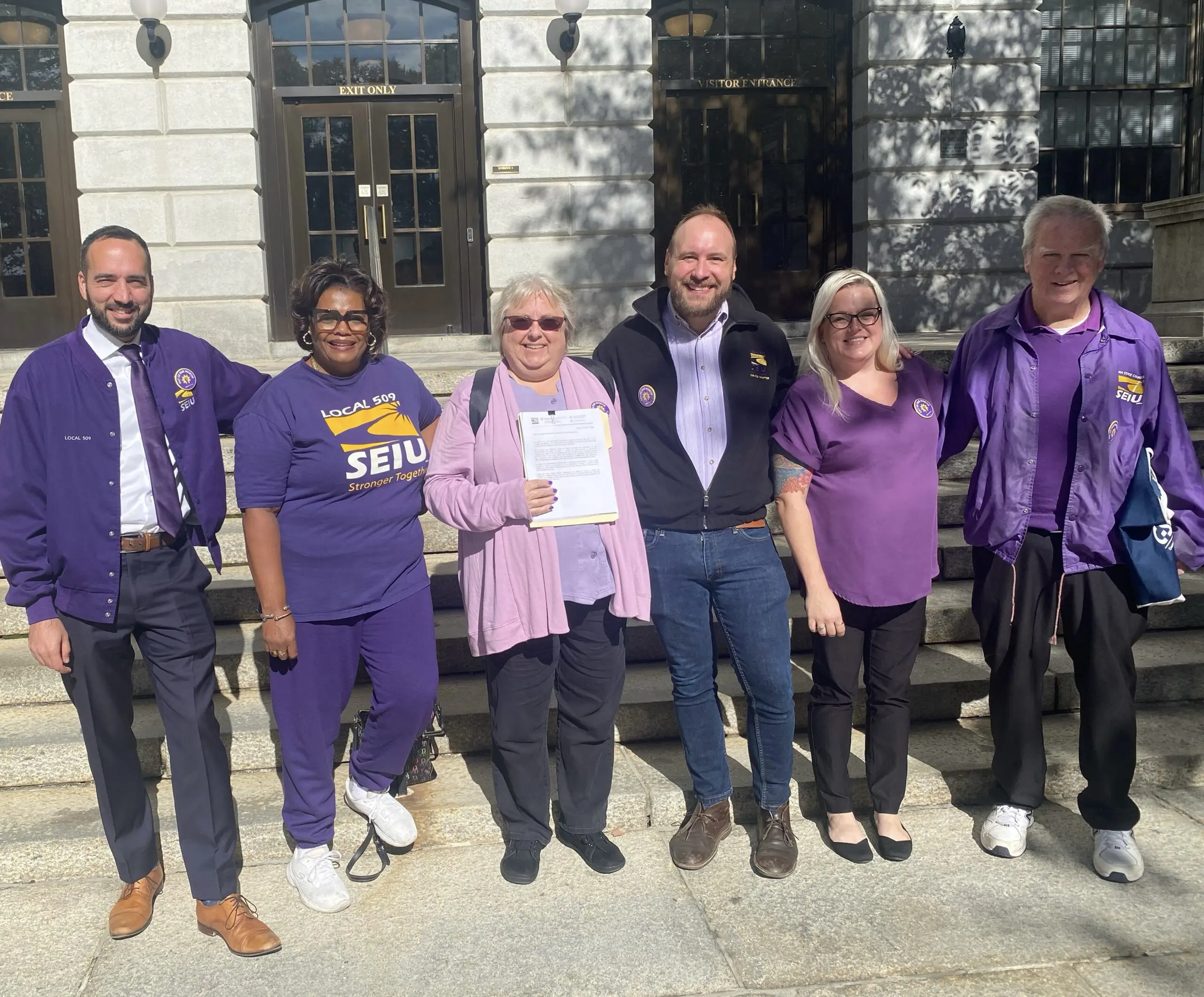 Six people stand outside the Massachusetts State House, smiling at the camera. Most are wearing purple SEIU clothing. The person in the center holds a document. Sunlight casts shadows on the building behind them.