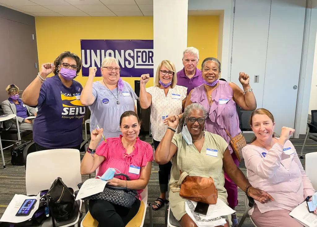 A diverse group of people, some wearing purple and union stickers, sit and stand together indoors, smiling and raising their fists in solidarity in front of a "UNIONS FOR ALL" sign.