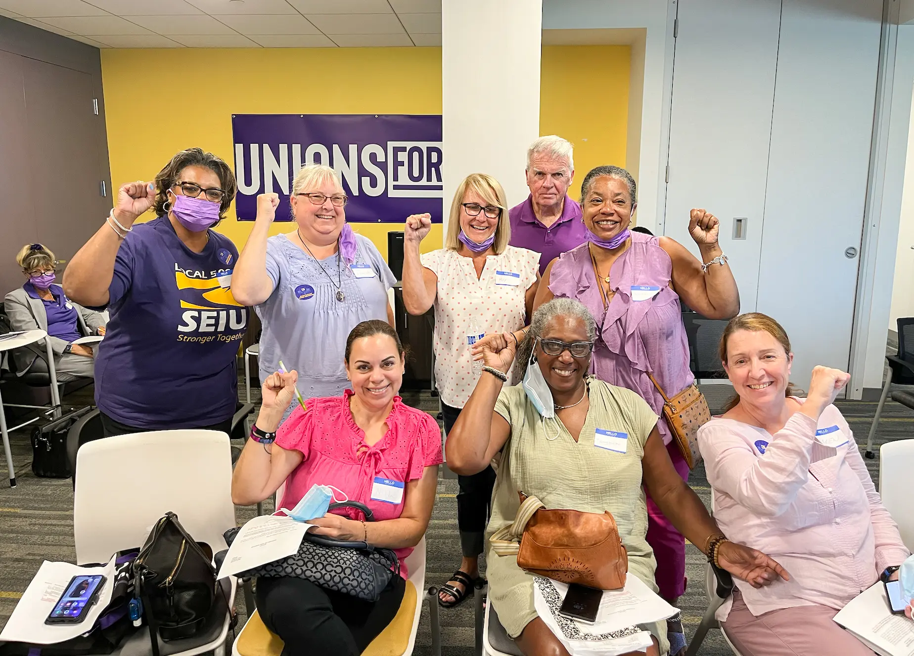 A diverse group of people, some wearing purple and union stickers, sit and stand together indoors, smiling and raising their fists in solidarity in front of a "UNIONS FOR ALL" sign.