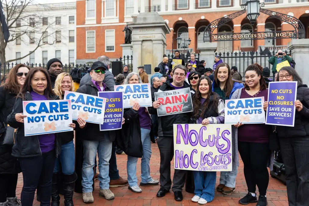 A diverse group of people stand outside the Massachusetts State House holding signs that read “Care Not Cuts,” “No Cuts No Closures,” and “No Cuts No Closures Save Our Services.”.