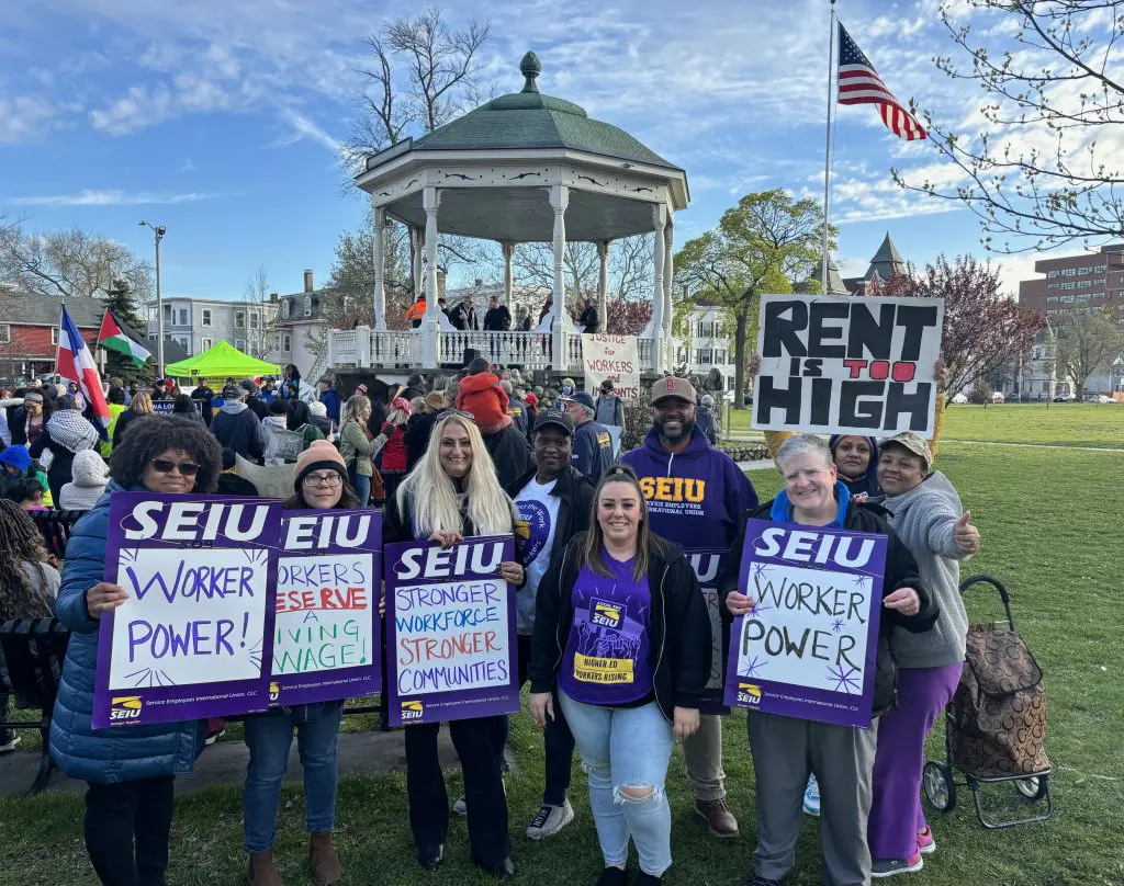 A group of people holding SEIU signs supporting worker power stands in front of a gazebo at a rally. Other people and signs, including one reading “RENT IS TOO HIGH,” are visible in the background. An American flag waves nearby.