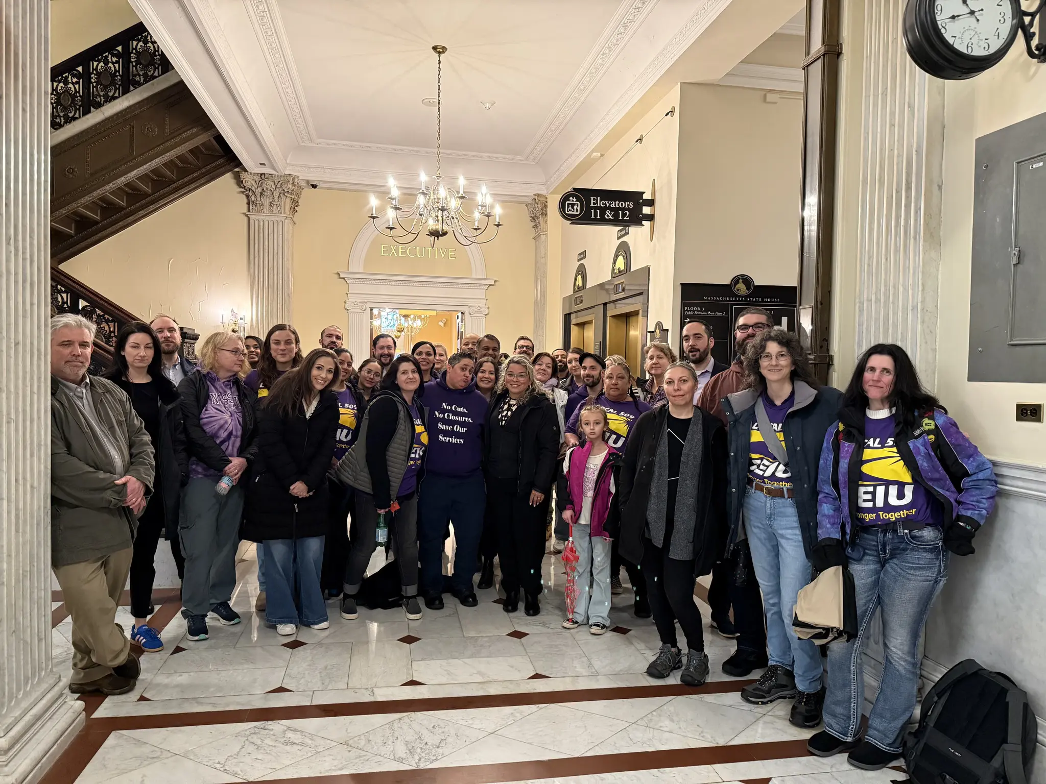 A large group of people poses together in the Massachusetts State House, some wearing purple SEIU shirts and holding signs. The setting features marble floors, columns, and chandeliers, with elevators and signage visible in the background.
