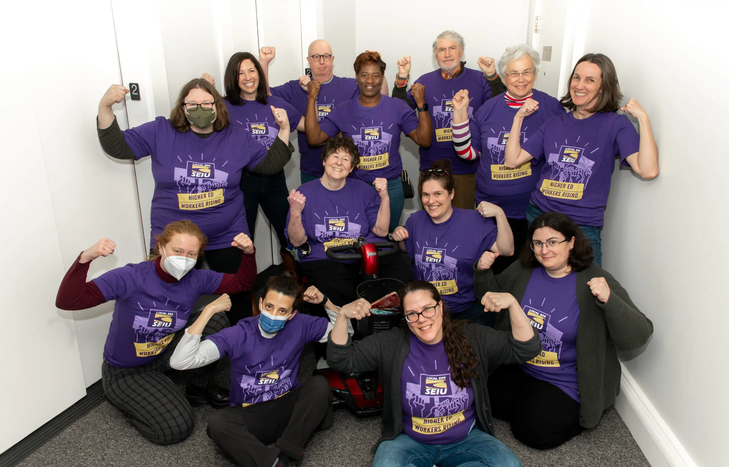 A group of thirteen people wearing matching purple SEIU t-shirts pose together indoors, smiling and flexing their arms in a triumphant gesture, showing solidarity.