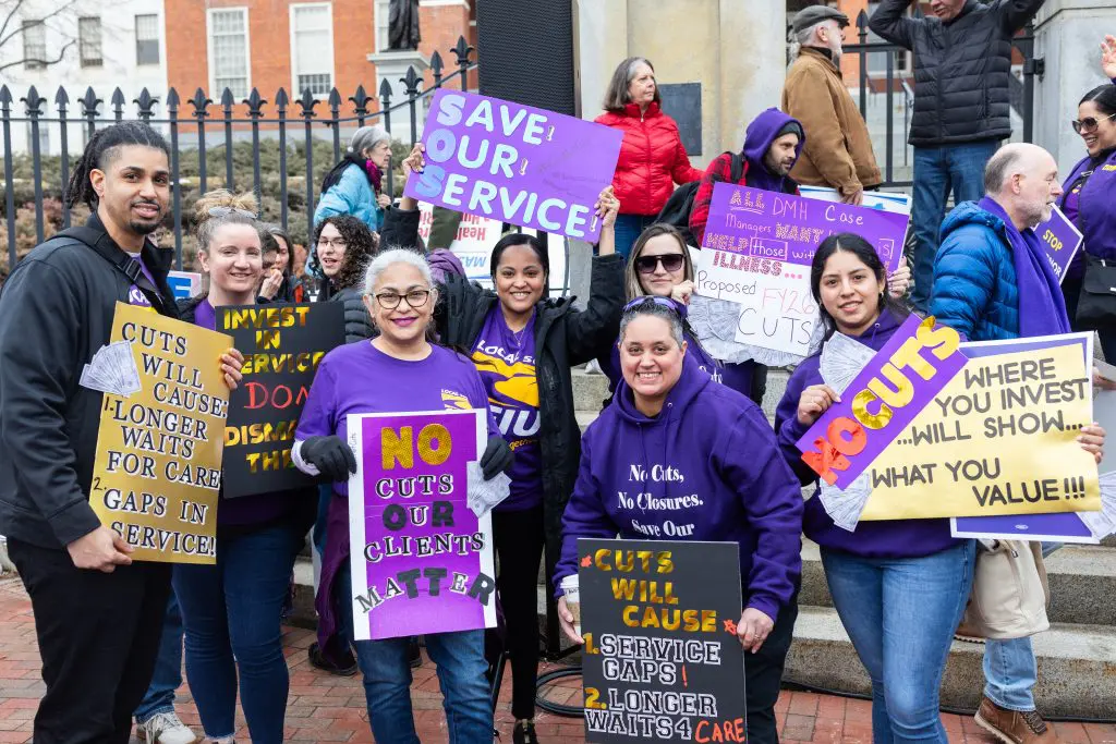 A group of people wearing purple hold colorful protest signs about service cuts and supporting workers. They are standing together and smiling at a rally in front of a building with an iron fence.