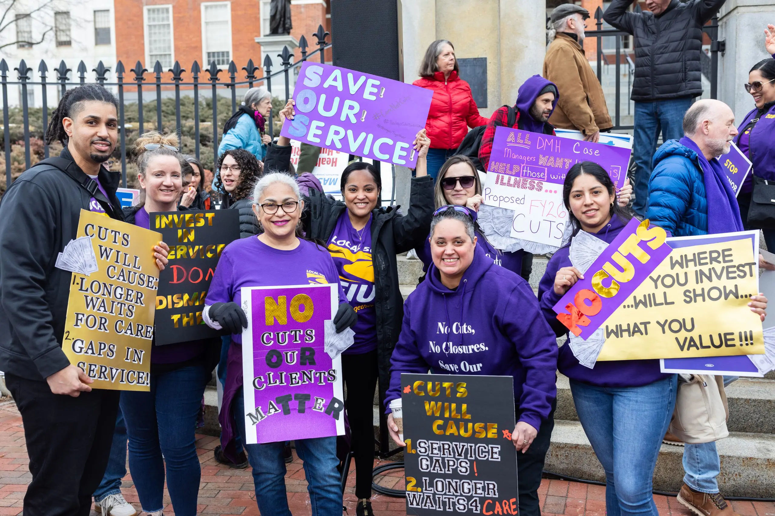 A group of people wearing purple hold colorful protest signs about service cuts and supporting workers. They are standing together and smiling at a rally in front of a building with an iron fence.