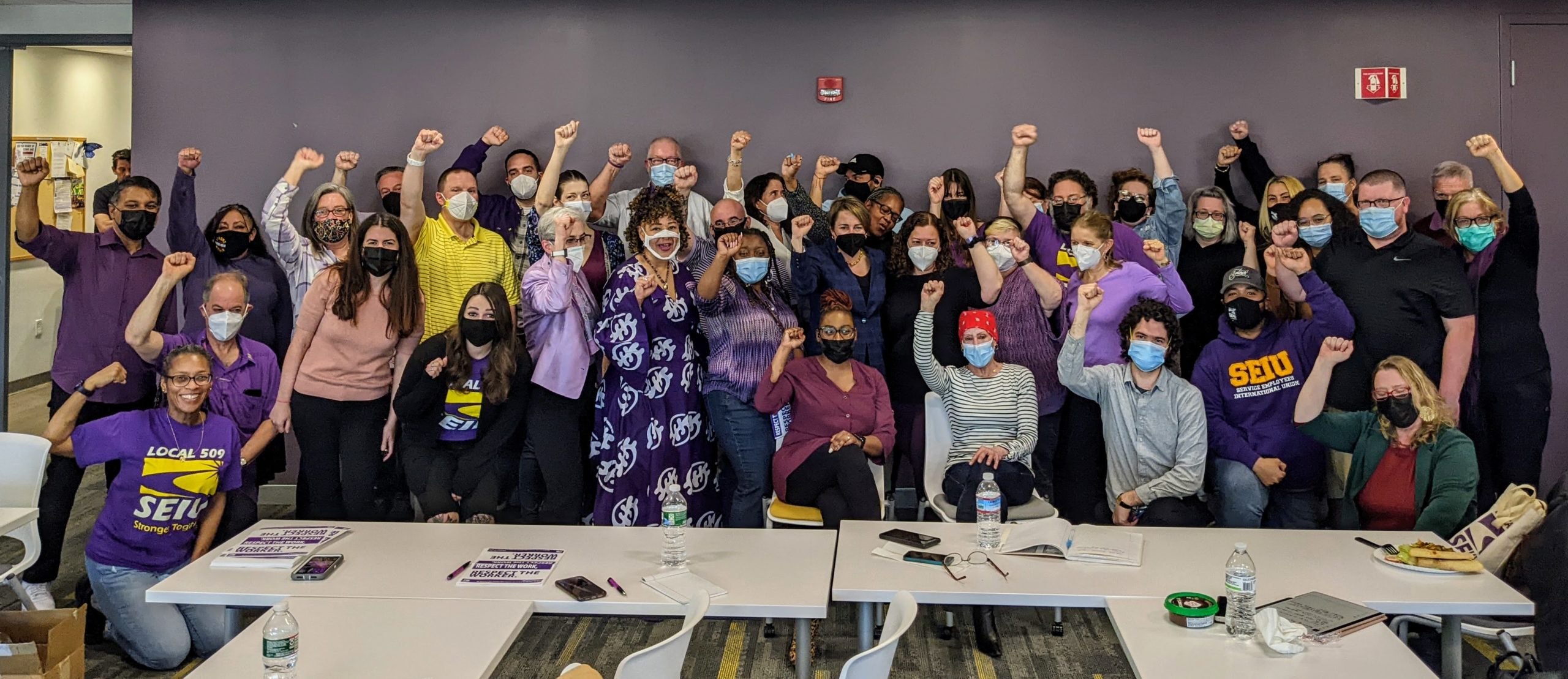 A large, diverse group of people pose indoors with raised fists, many wearing purple or SEIU shirts and face masks.