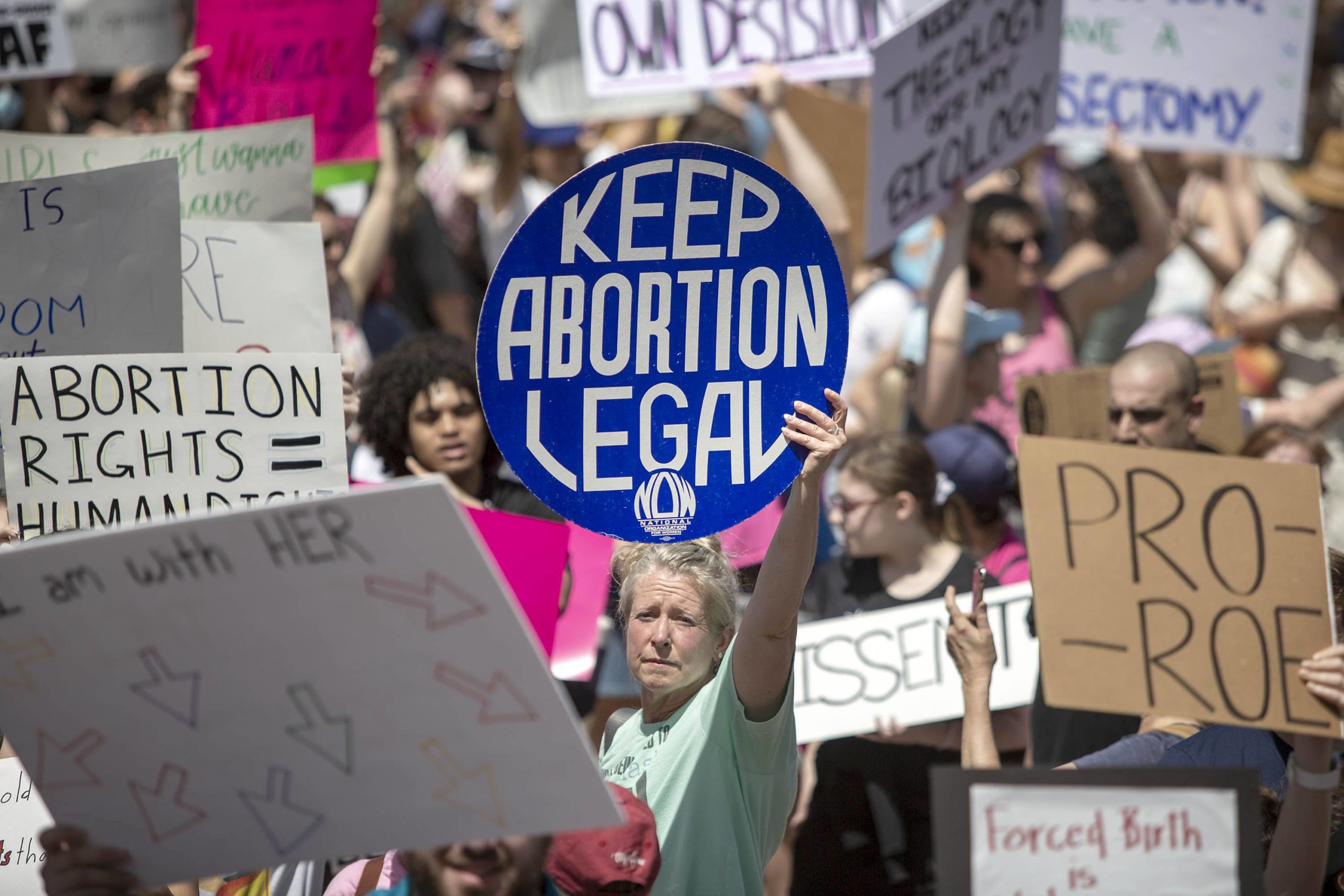 A crowd of protesters holds signs at a rally. A person in the center holds a large blue sign that reads "KEEP ABORTION LEGAL." Other signs with various messages about abortion rights are visible around them.