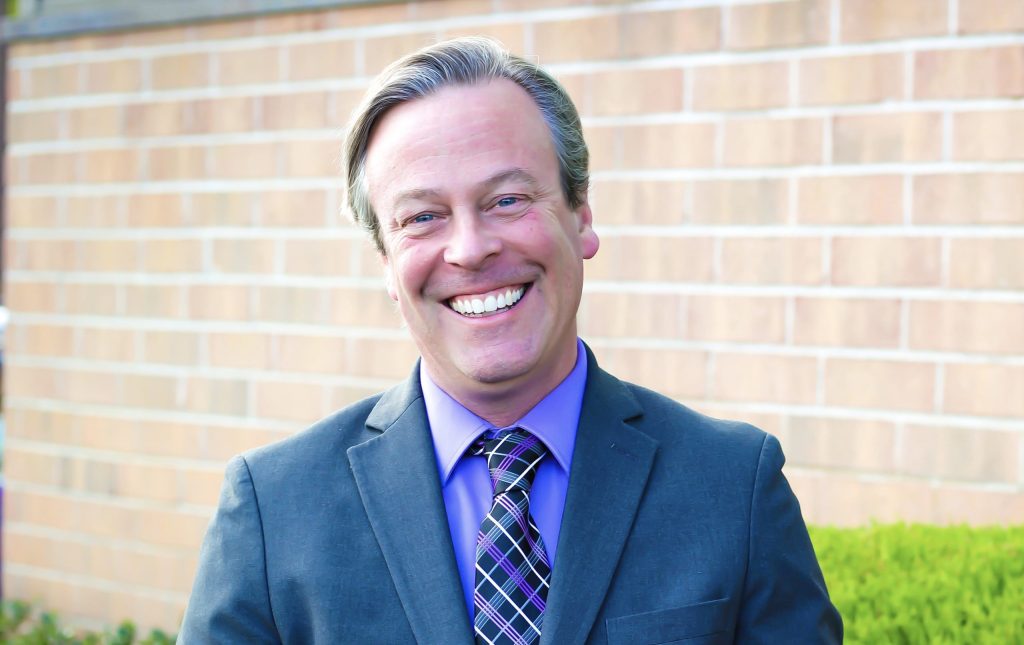 A man in a gray suit, purple shirt, and plaid tie smiles while standing outdoors in front of a brick wall and greenery.