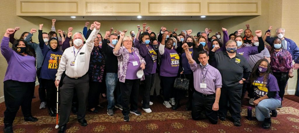 A large, diverse group of people, many wearing purple SEIU shirts, stand indoors with raised fists, showing solidarity.