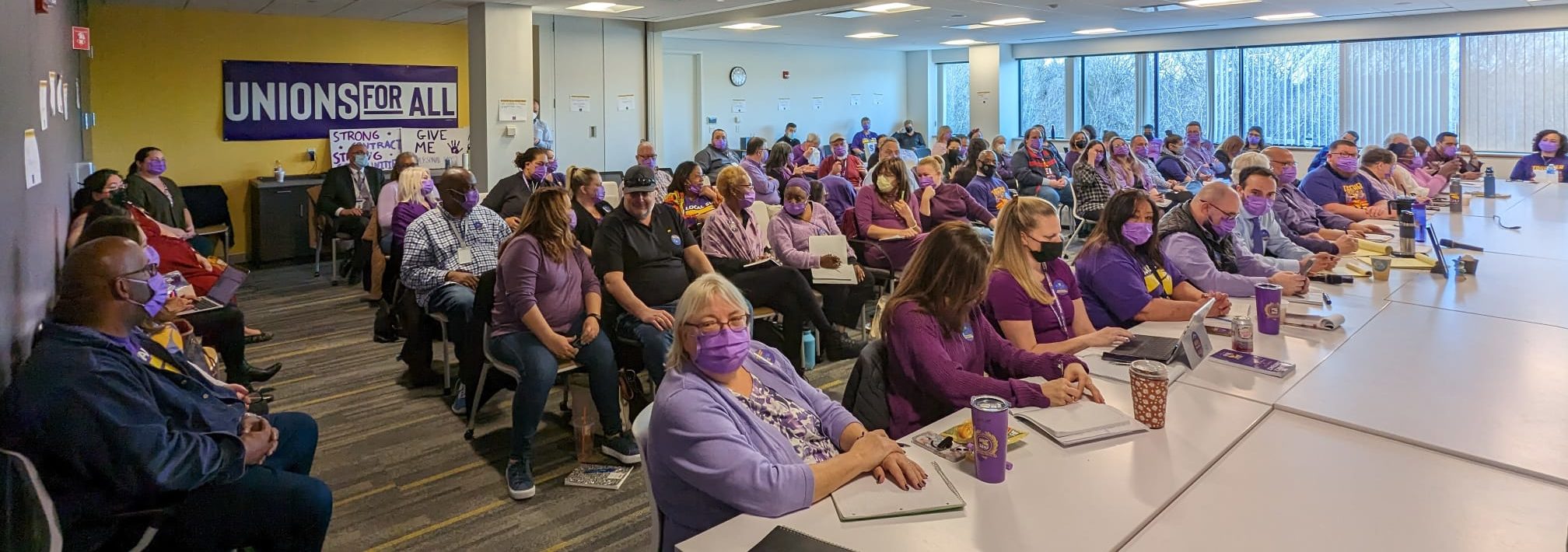 A large group of people, many wearing purple and some with masks, sit in a conference room with “UNIONS FOR ALL” and protest signs on the wall, attentively listening to a presentation.