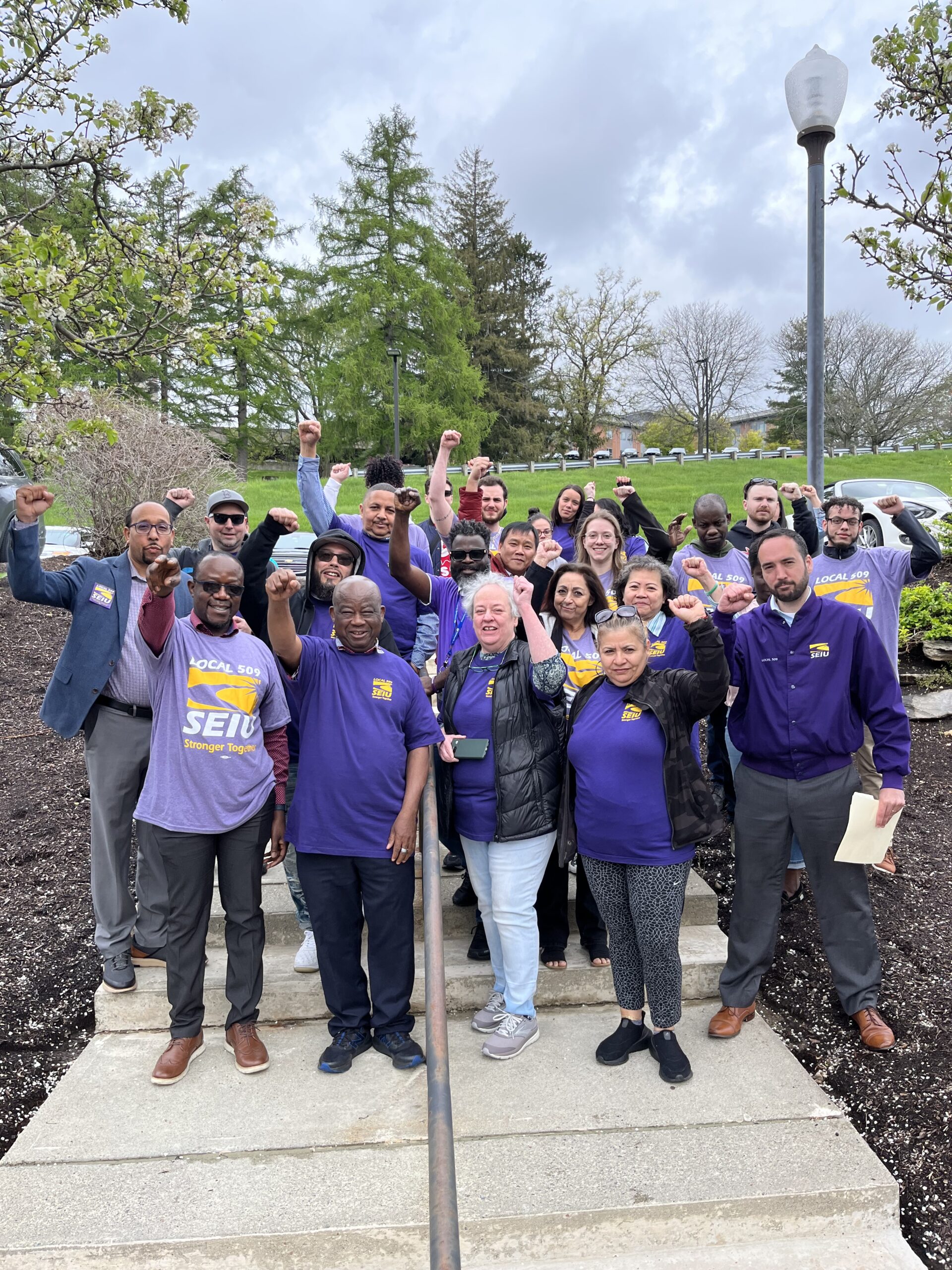 A diverse group of people in purple SEIU shirts stand outdoors, raising their fists in solidarity and smiling at the camera. Trees and cloudy skies are visible in the background.
