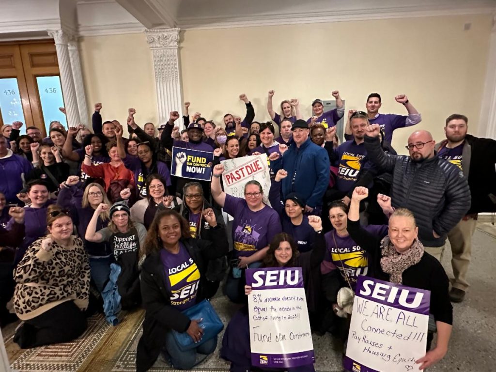 A large group of people, many wearing purple SEIU shirts, stand and kneel with raised fists, holding signs advocating for union support and funding, inside the Massachusetts State House.