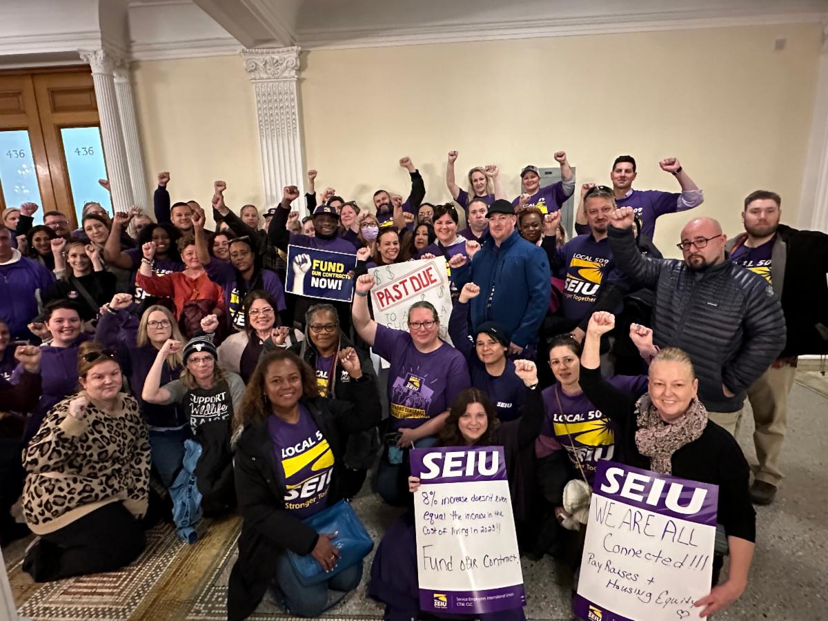 A large group of people, many wearing purple SEIU shirts, stand and kneel with raised fists, holding signs advocating for union support and funding, inside the Massachusetts State House.