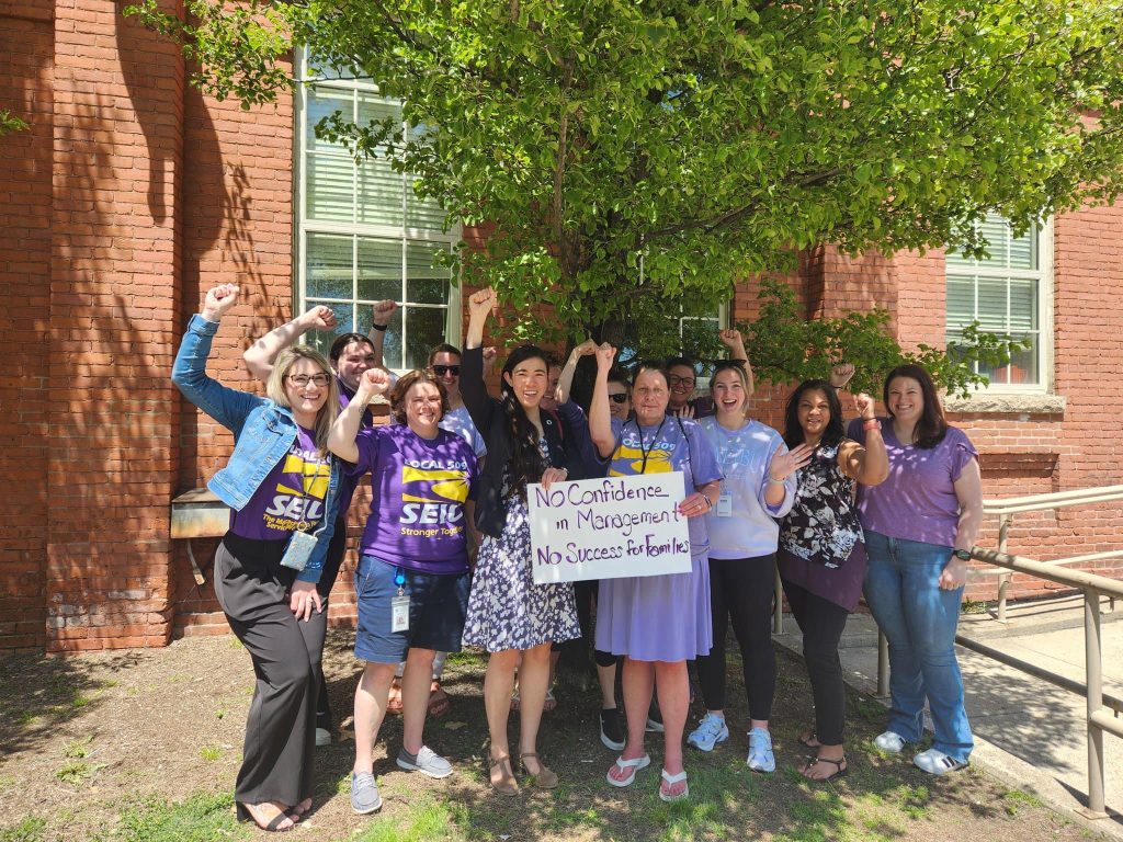 A group of people stand outside a brick building, some raising fists in solidarity. Two people in the center hold a sign reading, "No Confidence in Management, No Success for Families!"