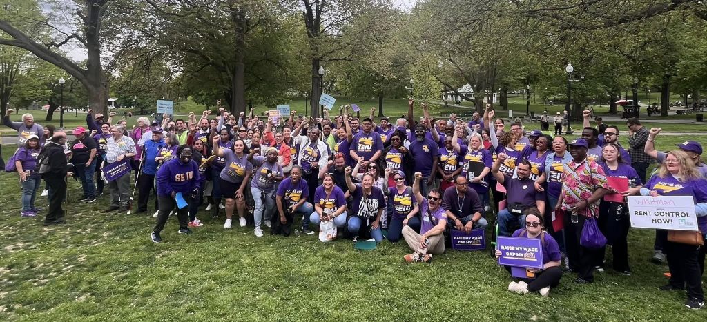 A large group of people wearing purple shirts and holding signs gather in a park, raising their fists in solidarity for rent control and fair wages. Trees and green grass are visible in the background.