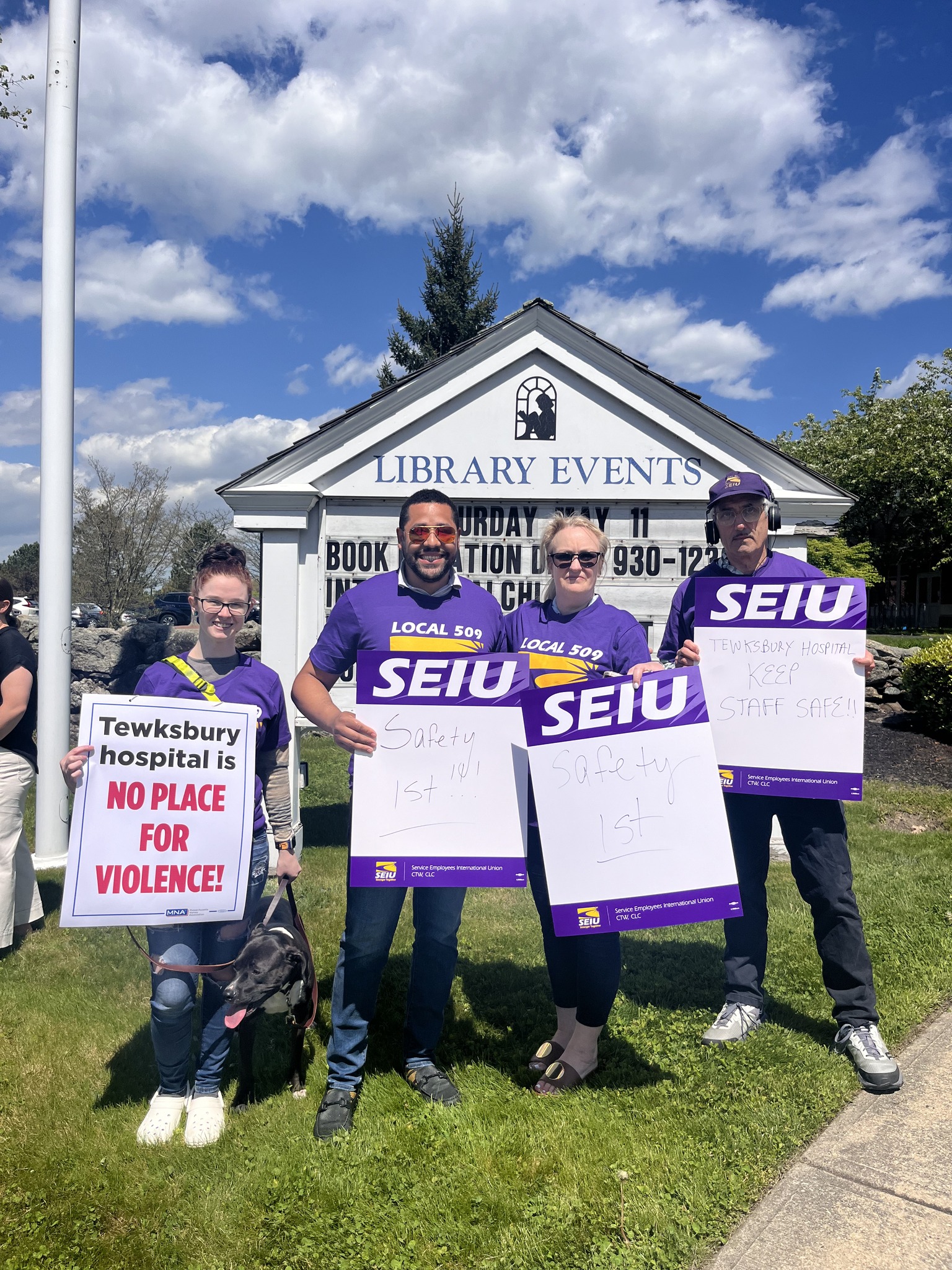 Four people stand outside holding protest signs in front of a "Library Events" board. Three hold purple SEIU signs advocating for safety, while one holds a sign reading "Tewksbury hospital is NO PLACE FOR VIOLENCE!.