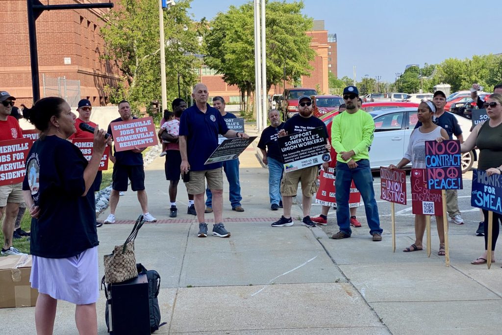A group of people stand outside holding signs that support union workers and protest against union busting. One woman speaks to the group while others listen attentively.
