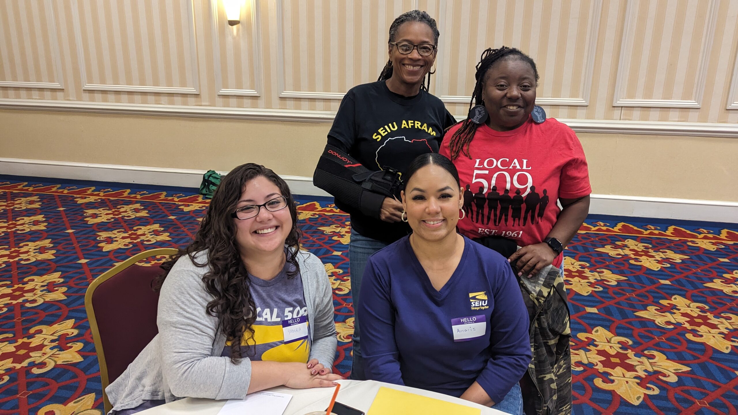 Four women smile at the camera during a meeting or event. Two are seated at a table with papers, while two stand behind them. They wear shirts with "Local 509" and "SEIU Afram" logos. The setting is a carpeted, banquet-style room.