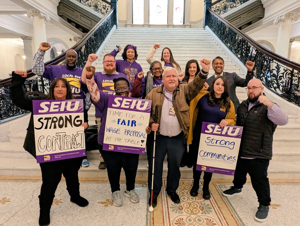A diverse group of people stand on marble stairs, holding SEIU signs reading “STRONG CONTRACT,” “TIME FOR A FAIR WAGE PROPOSAL AT THE TABLE,” and “STRONG COMMUNITIES.” Most wear purple shirts and raise fists in solidarity.