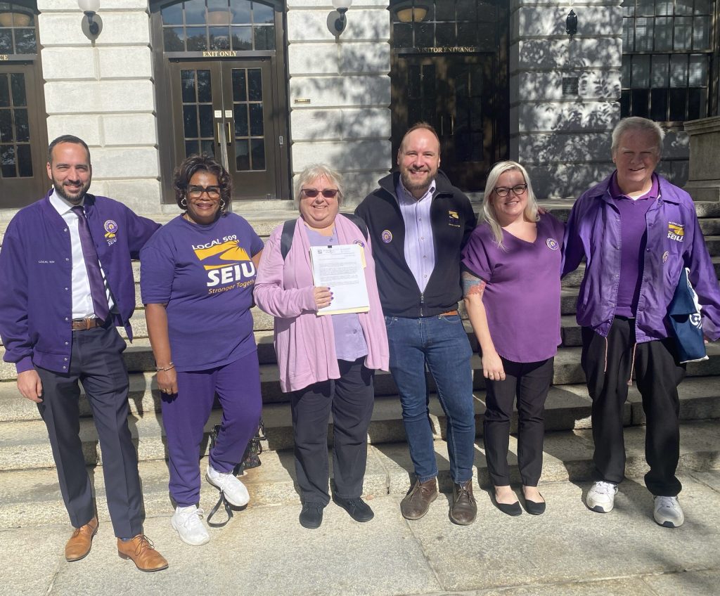 Six people stand outside the Massachusetts State House, smiling at the camera. Most are wearing purple SEIU clothing. The person in the center holds a document. Sunlight casts shadows on the building behind them.