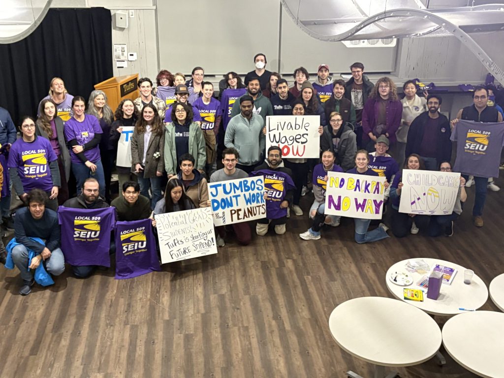 A large group of people poses indoors, many wearing purple SEIU shirts and holding signs with messages like “Livable Wages Now,” “Childcare Now,” and “End Backpay Now.”.