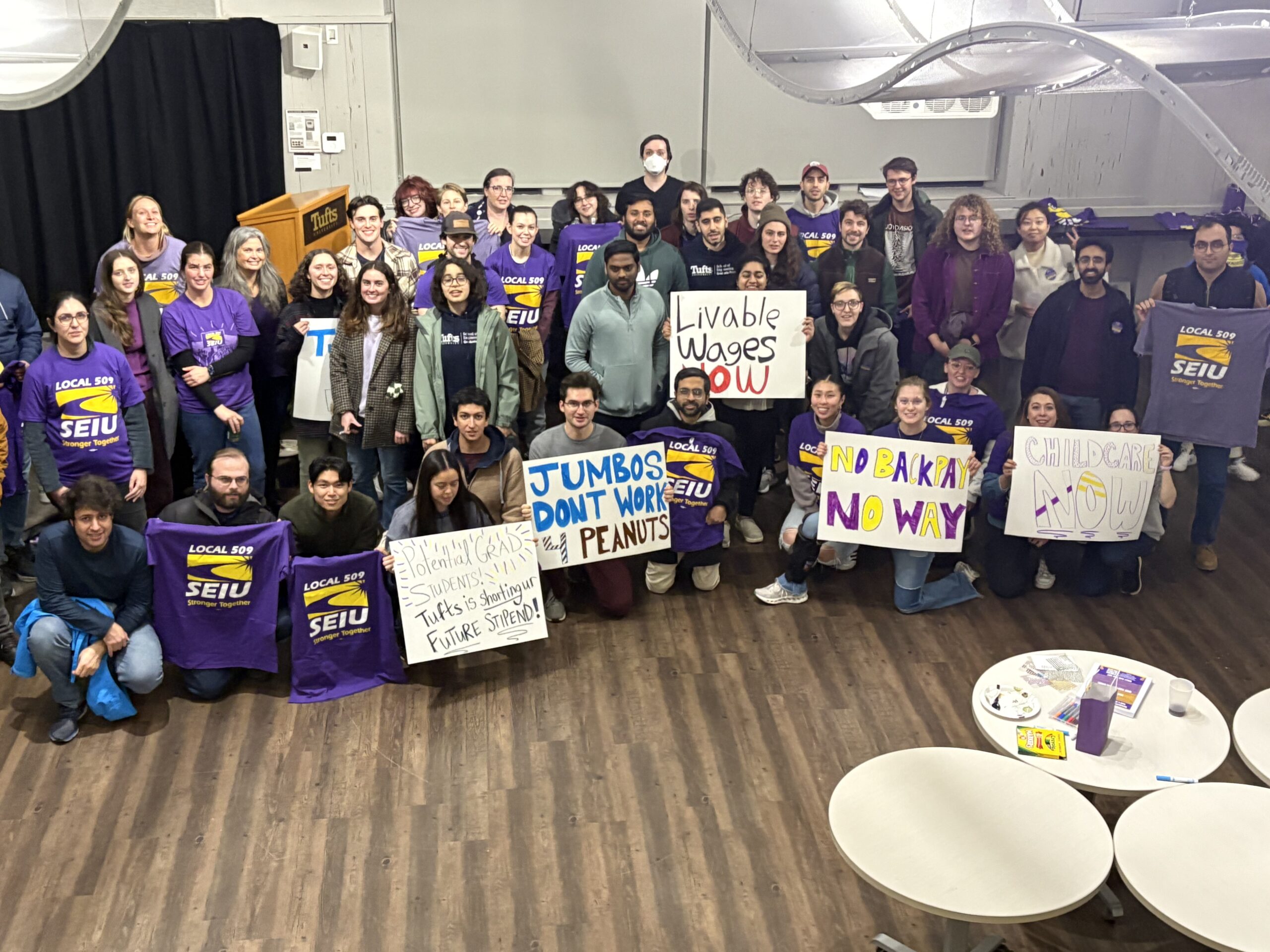 A large group of people poses indoors, many wearing purple SEIU shirts and holding signs with messages like “Livable Wages Now,” “Childcare Now,” and “End Backpay Now.”.