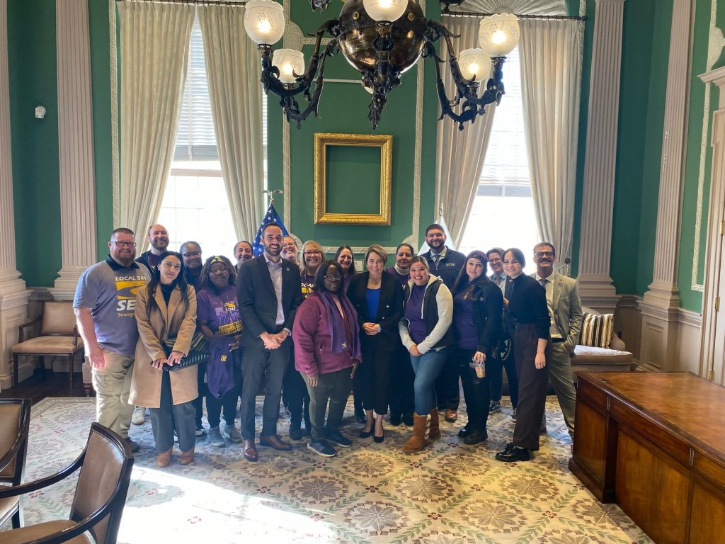 A group of people stand together and smile for a photo with Governor Maura Healey in a formal room with green walls, large windows, and an American flag in the background. Several people wear purple shirts with SEIU logos.