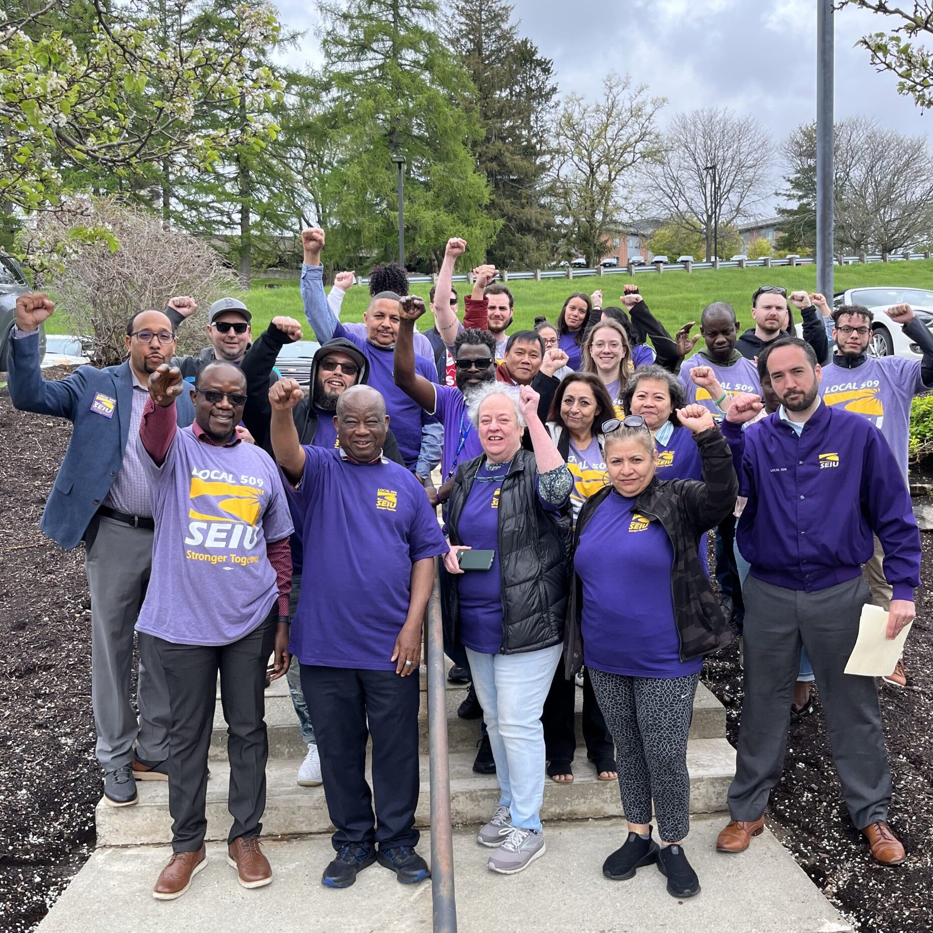 A diverse group of people in purple SEIU shirts stand outdoors, raising their fists in solidarity and smiling at the camera. Trees and cloudy skies are visible in the background.