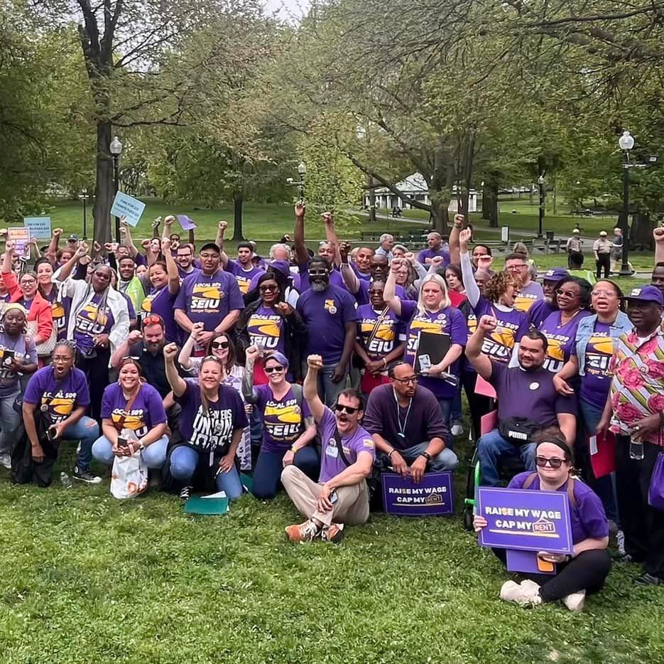 A large group of people wearing purple shirts and holding signs gather in a park, raising their fists in solidarity for rent control and fair wages. Trees and green grass are visible in the background.