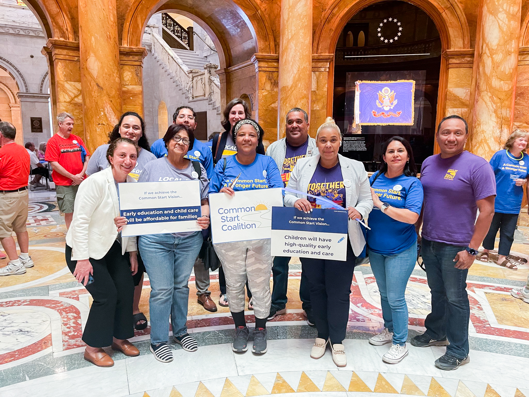 A diverse group of people stands in a grand marble building holding signs supporting affordable, high-quality early education and child care. They are smiling and wearing shirts with advocacy messages.
