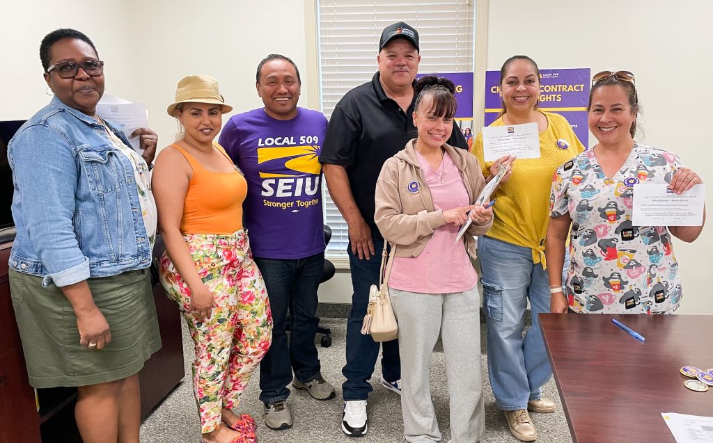A group of seven people, some holding papers, stand together smiling in an office with SEIU Local 509 union posters visible on the wall behind them.