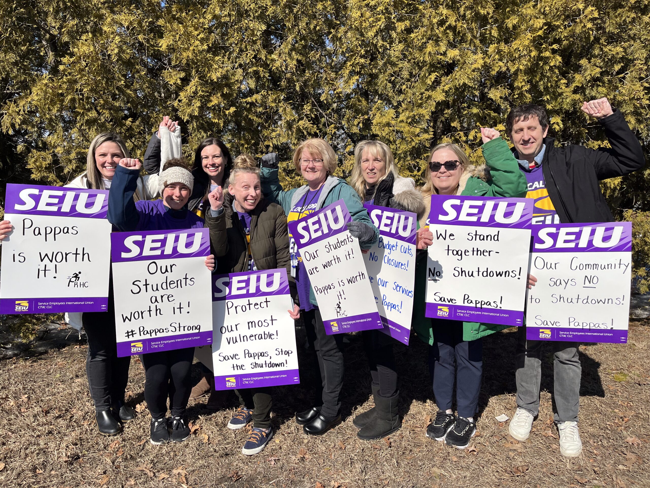 A group of people stand outdoors holding SEIU signs that read messages like “Pappas is worth it!,” “Our Students are worth it!,” and “Save Pappas!” They are smiling and raising their fists in solidarity.