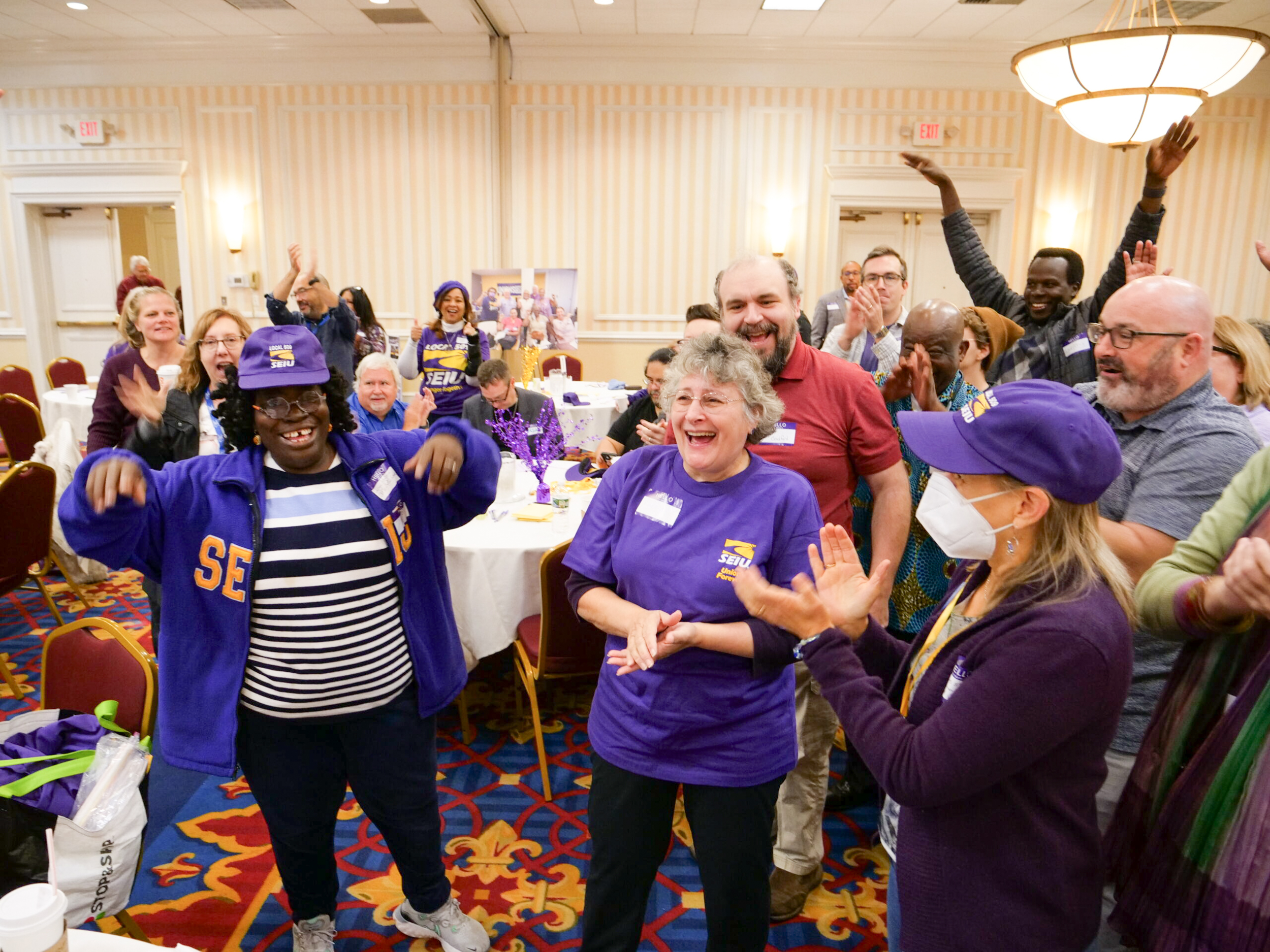 A diverse group of people stands in a brightly lit room, smiling, clapping, and cheering. Most are wearing purple SEIU shirts and hats, showing excitement and camaraderie at a lively event or meeting.