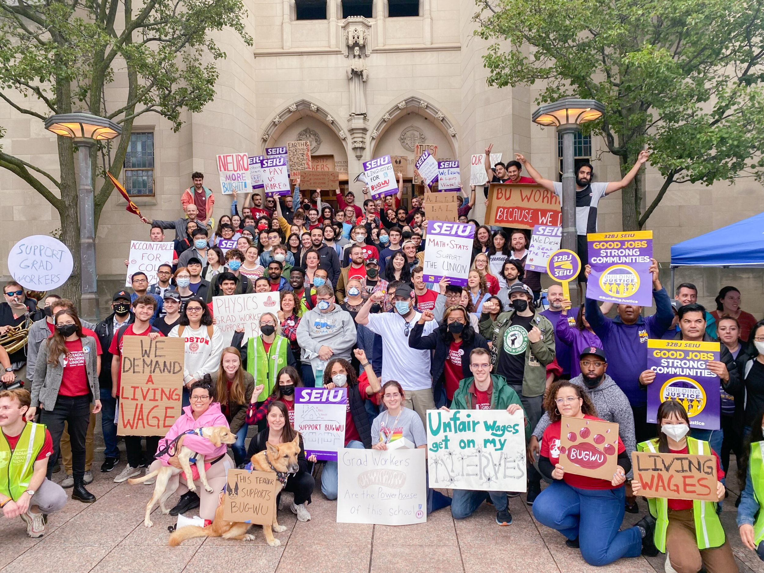 A large group of people stand in front of a stone building, holding signs with messages such as “Living Wages,” and “Good Jobs, Strong Communities,” rallying for workers’ rights and fair pay.