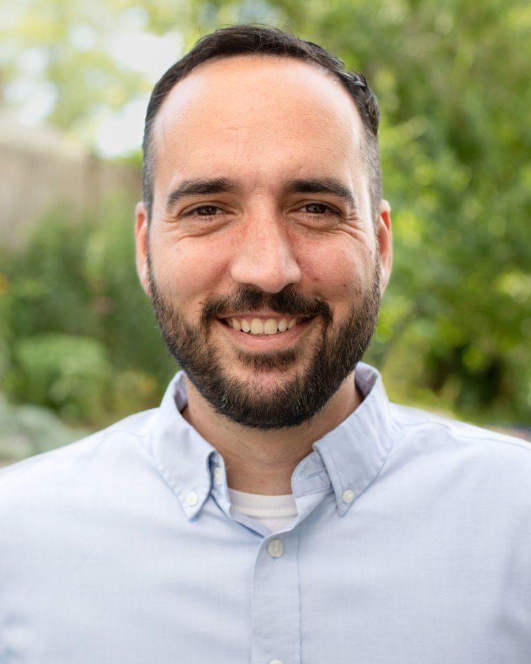 A man with short dark hair and a beard, wearing a light blue button-up shirt, smiles at the camera outdoors with greenery blurred in the background.