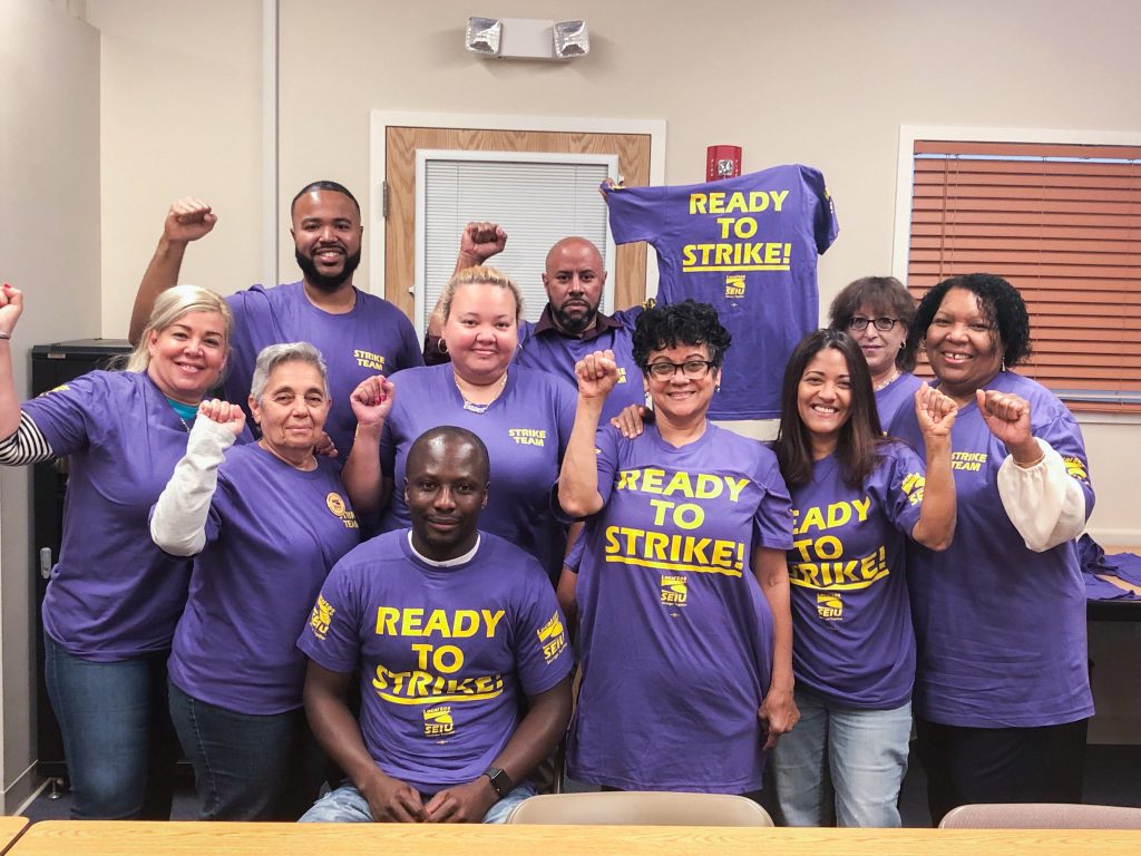 Ten people wearing matching purple "Ready to Strike!" shirts stand and sit together indoors, smiling and raising their fists in solidarity. A similar shirt is hung on a door in the background.