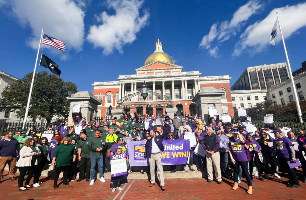 A large group of people gathers in front of the Massachusetts State House, holding SEIU banners and wearing purple shirts.
