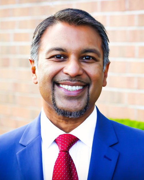 A man in a blue suit, white shirt, and red tie is smiling in front of a brick wall background. He has short dark hair, a trimmed beard, and appears to be outdoors.