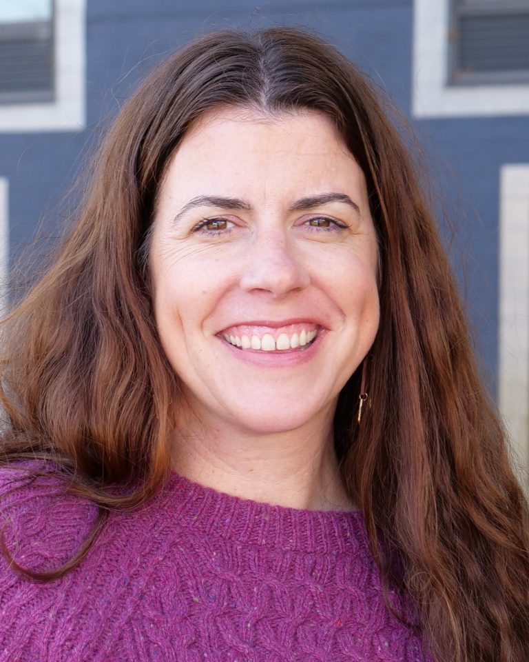 A woman with long brown hair, wearing a textured purple sweater, smiles at the camera. The background shows a dark-colored building with windows.